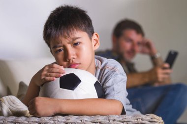 young sad and bored child at home couch feeling frustrated and unattended waiting his father for playing football while man networking on mobile phone as internet addict father neglecting son