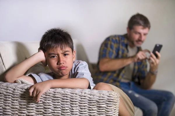young sad and bored 7 or 8 years old child at home couch feeling frustrated and unattended while man networking on mobile phone as internet addict father neglecting his young son