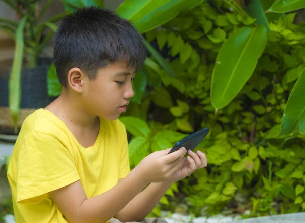 isolated lifestyle portrait of 7 or 8 years old Asian child focused and concentrated playing with mobile phone outdoors at home garden in kid suffering gaming addiction concept