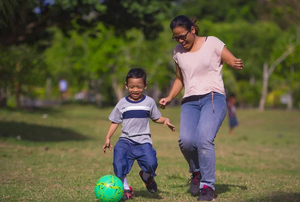 portrait in city park of happy Asian Indonesian mother playing football with little 5 years old son running together excited laughing having fun in soccer fan child and healthy lifestyle education