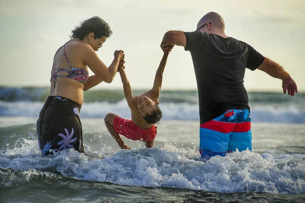 young happy mixed ethnicity couple Caucasian man and Asian Indonesian woman playing at tropical beach with little son the parents raising up the child above the sea water having fun in holidays trip