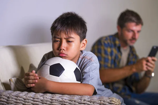 young sad and bored child at home couch feeling frustrated and unattended waiting his father for playing football while man networking on mobile phone as internet addict father neglecting son