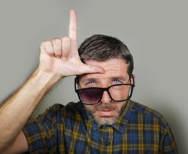 30s or 40s unshaven sad and ashamed man in weird broken nerdy glasses doing loser sign with hand and fingers on his forehead with funny depressed expression in unsuccessful geek concept