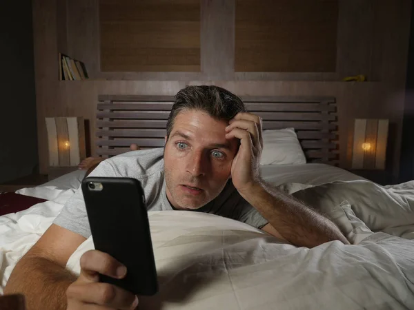 young attractive surprised and shocked man using mobile phone at home lying on bed late at night in surprise face expression while networking or using social media in internet addiction concept