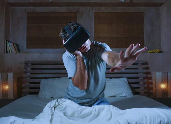 young excited and amazed man wearing VR virtual reality goggles device playing internet 3D simulator game enjoying exciting and shocking visual experience at home bed