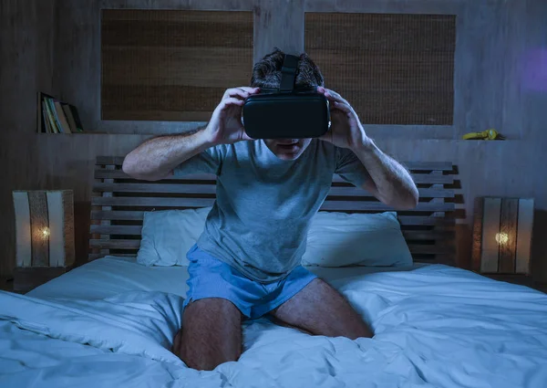 young excited and amazed man wearing VR virtual reality goggles device playing internet 3D simulator game enjoying exciting and shocking visual experience at home bed