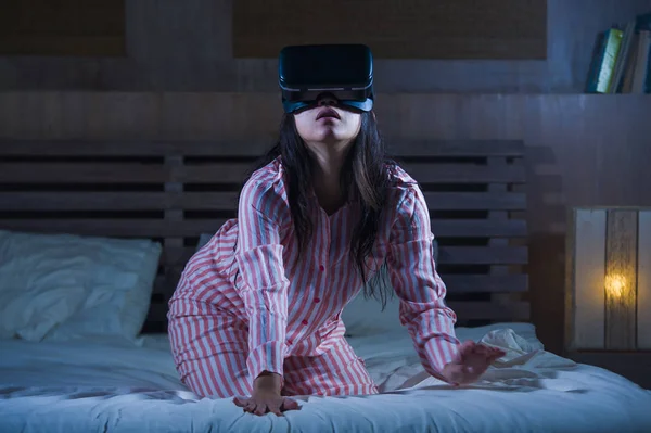 young happy and excited girl playing with virtual reality VR goggles headset device having fun on bed having 3D experience with video game shocked looking and touching digital illusion