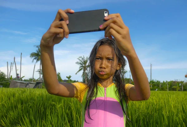 7 or 8 years old sweet and pretty female child outdoors at rice field landscape taking selfie portrait  photo with mobile phone camera enjoying holidays in children and internet technology concept