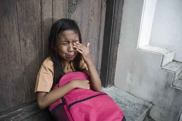 7 oder 8 Jahre Kind in Schuluniform, das draußen auf dem Boden sitzt und weint, traurig und deprimiert ihren Rucksack haltend, das Mobbing- und Missbrauchsproblem fühlend allein und hilflos — Stockfoto