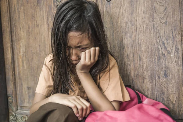 7 oder 8 Jahre Kind in Schuluniform, das draußen auf dem Boden sitzt und weint, traurig und deprimiert ihren Rucksack hält und unter Mobbing- und Missbrauchsproblemen leidet, fühlt sich hilflos — Stockfoto