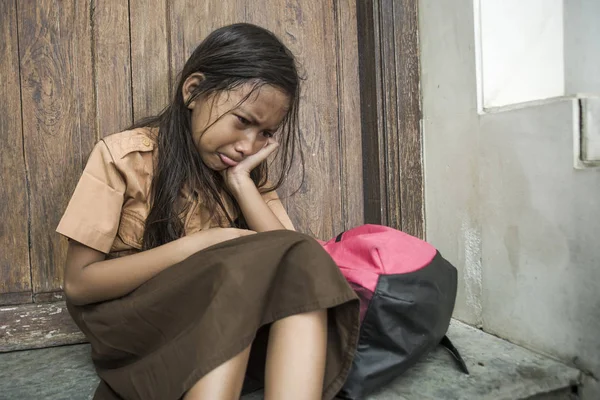 7 oder 8 Jahre Kind in Schuluniform, das draußen auf dem Boden sitzt und weint, traurig und deprimiert ihren Rucksack hält und unter Mobbing- und Missbrauchsproblemen leidet, fühlt sich hilflos — Stockfoto