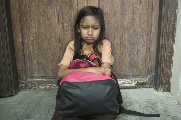 7 oder 8 Jahre Kind in Schuluniform, das draußen auf dem Boden sitzt und weint, traurig und deprimiert ihren Rucksack hält und unter Mobbing- und Missbrauchsproblemen leidet, fühlt sich hilflos — Stockfoto