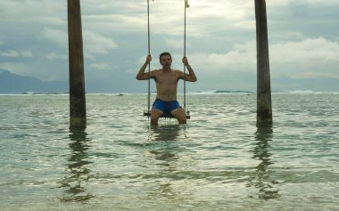 young fit and attractive man enjoying Summer holidays swinging carefree on sea swing at tropical island beach resort having fun relaxed in amazing travel tourist destination concept clipart
