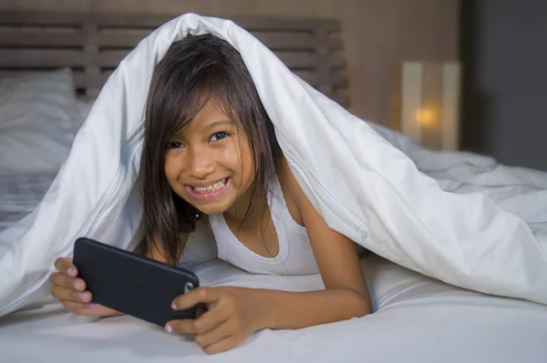 Feliz y hermoso niño de 7 años de edad que se divierten jugando juego de Internet con el teléfono móvil acostado en la cama alegre y emocionado en la niña y el concepto de tecnología — Foto de Stock