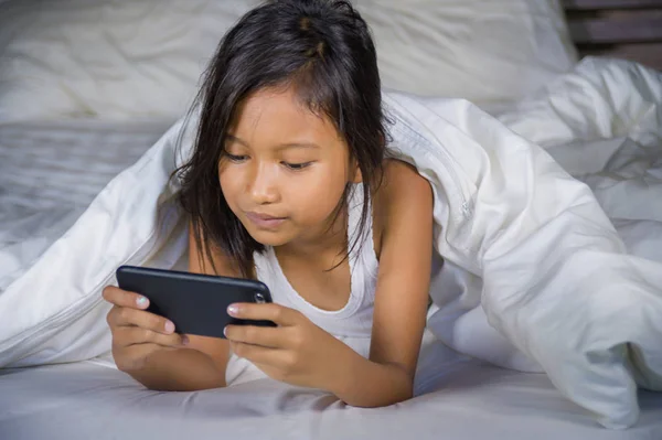 Feliz y hermoso niño de 7 años de edad que se divierten jugando juego de Internet con el teléfono móvil acostado en la cama alegre y emocionado en la niña y el concepto de tecnología — Foto de Stock