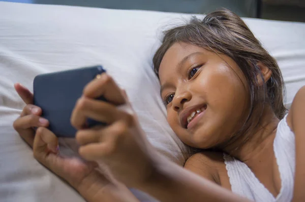 Feliz y hermoso niño de 7 años de edad que se divierten jugando juego de Internet con el teléfono móvil acostado en la cama alegre y emocionado en la niña y el concepto de tecnología — Foto de Stock