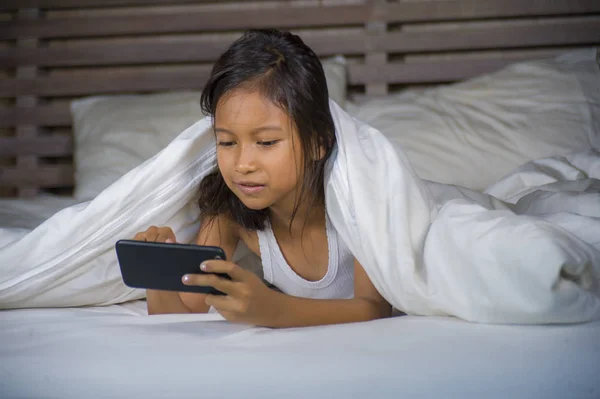 Feliz y hermoso niño de 7 años de edad que se divierten jugando juego de Internet con el teléfono móvil acostado en la cama alegre y emocionado en concepto de tecnología para niños y domésticos — Foto de Stock