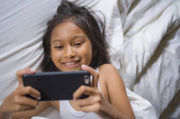 Feliz y hermoso niño de 7 años de edad que se divierten jugando juego de Internet con el teléfono móvil acostado en la cama alegre y emocionado en concepto de tecnología para niños y domésticos — Foto de Stock