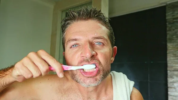 lifestyle portrait of young handsome and attractive man with blue eyes at home bathroom with towel on shoulder washing teeth with toothbrush in dental hygiene