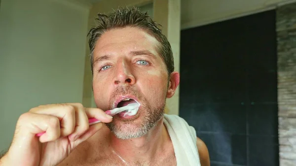 lifestyle portrait of young handsome and attractive man with blue eyes at home bathroom with towel on shoulder washing teeth with toothbrush in dental hygiene