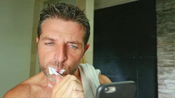 Retrato de joven apuesto y atractivo adicto a Internet hombre en el baño en casa con toalla en el hombro lavado de dientes con cepillo de dientes utilizando el teléfono móvil en línea — Foto de Stock