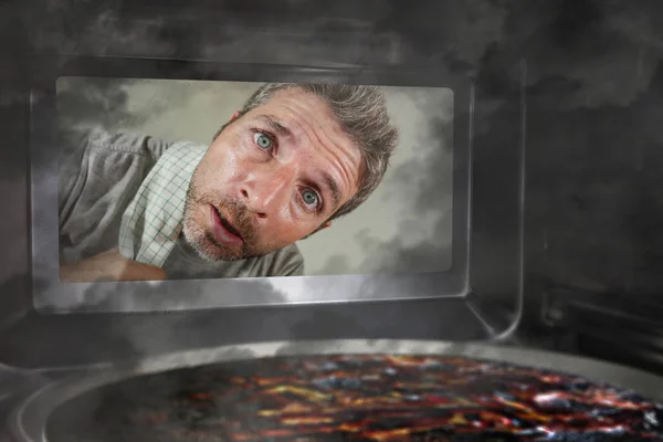 Messy and funny dummy man in the kitchen looking through microwave or oven pizza burning overcooked making a mess of home cook in domestic disaster and lifestyle concept — Stock Photo, Image