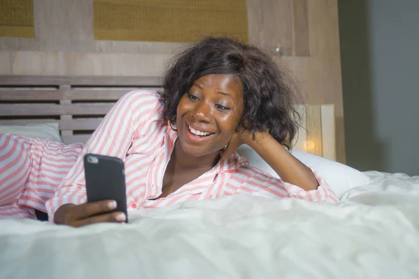 Joven feliz y hermosa mujer afroamericana negra en pijama acostado relajado sonriente alegre y fresco usando internet teléfono móvil en red o en línea citas —  Fotos de Stock