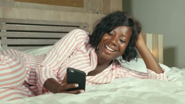 美しく 幸せな黒いアフリカ系アメリカ人の若い女性パジャマ リラックスして居心地の良いインターネット携帯電話オンライン デートまたは陽気な笑みを浮かべてソーシャル メディアを楽しむとのネットワー キングのベッドで横になっています — ストック動画