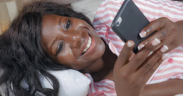 Joven hermosa y feliz negro afro mujer americana en pijama tumbado relajado en la cama en red con internet teléfono móvil en línea citas o disfrutar de las redes sociales —  Fotos de Stock