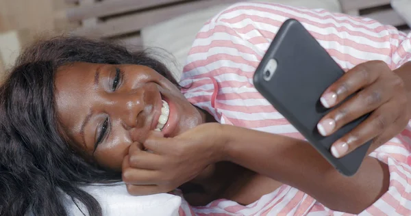 Молодая красивая и счастливая чернокожая афро-американка в пижаме лежала расслабленной на кровати, общаясь с интернет-телефоном онлайн знакомства или наслаждаясь социальными сетями — стоковое фото