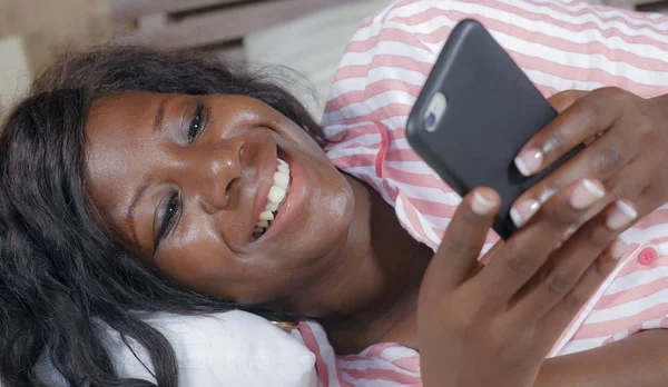 Young beautiful and happy black afro American woman in pajamas lying relaxed on bed networking with internet mobile phone online dating or enjoying social media — Stock Photo, Image