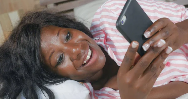 Joven hermosa y feliz negro afro mujer americana en pijama tumbado relajado en la cama en red con internet teléfono móvil en línea citas o disfrutar de las redes sociales —  Fotos de Stock