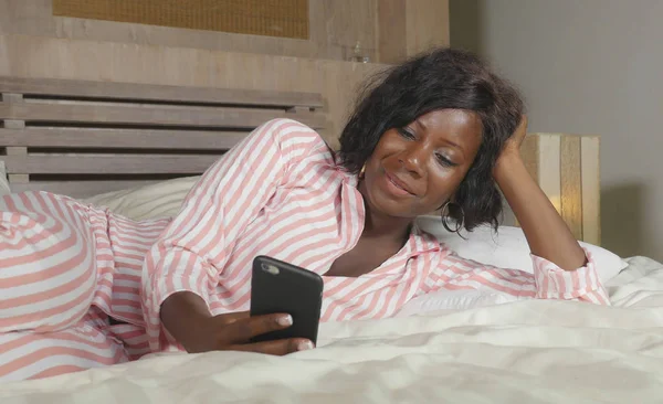 美しく、幸せな黒いアフロ アメリカンの若い女性インターネット携帯電話オンライン デートやソーシャル メディアを楽しむとのネットワー キングのベッドでリラックスして横になっているパジャマ — ストック写真