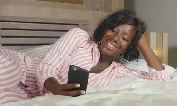 Joven hermosa y feliz negro afro mujer americana en pijama tumbado relajado en la cama en red con internet teléfono móvil en línea citas o disfrutar de las redes sociales — Foto de Stock