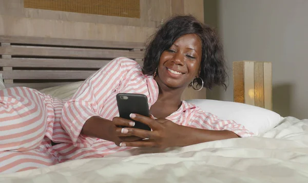 Молодая красивая и счастливая чернокожая афро-американка в пижаме лежала расслабленной на кровати, общаясь с интернет-телефоном онлайн знакомства или наслаждаясь социальными сетями — стоковое фото