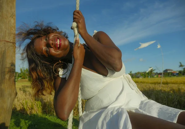 Jovem feliz e bonita negra afro americana em vestido de verão jogando ao ar livre no balanço sorrindo alegre e relaxado se divertindo balançando em fundo tropical — Fotografia de Stock