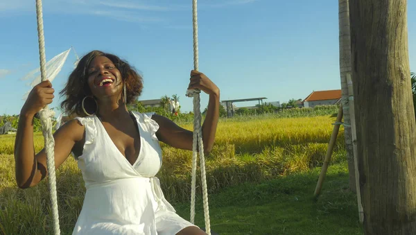 Jovem feliz e bonita negra afro americana em vestido de verão jogando ao ar livre no balanço sorrindo alegre e relaxado se divertindo balançando em fundo tropical — Fotografia de Stock