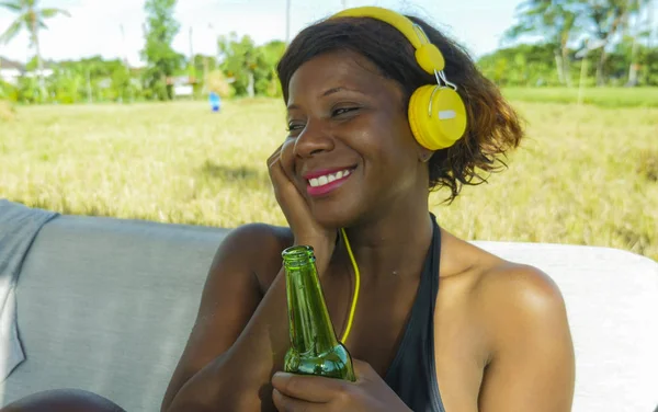 Beautiful and happy black afro American woman with headset listening to music outdoors chilled and relaxed enjoying summer holidays smiling cheerful drinking beer — Stock Photo, Image