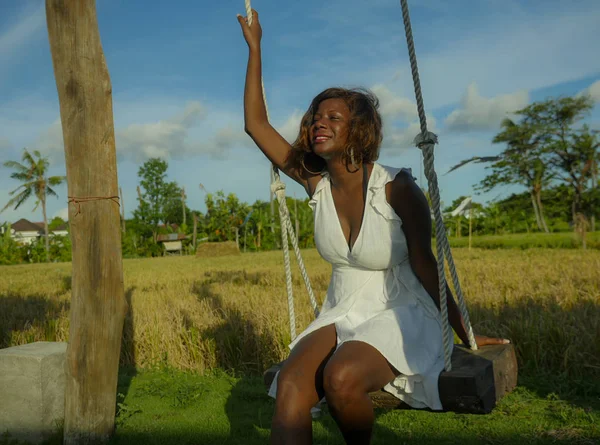 Jovem feliz e bonita negra afro-americana em vestido de verão jogando ao ar livre no balanço sorrindo alegre e relaxado se divertindo balançando em feriados tropicais — Fotografia de Stock