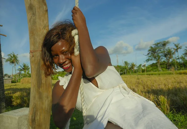 Jovem feliz e bonita negra afro-americana em vestido de verão jogando ao ar livre no balanço sorrindo alegre e relaxado se divertindo balançando em feriados tropicais — Fotografia de Stock