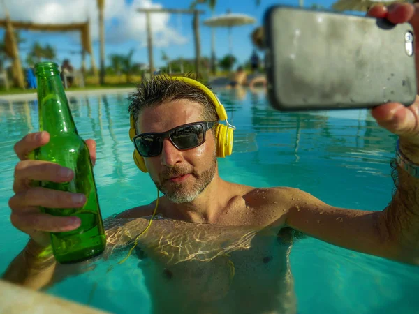 Joven feliz y atractivo hombre tomando foto selfie con el teléfono móvil bebiendo cerveza y escuchando música en la piscina tropical resort, piscina disfrutando de vacaciones de verano —  Fotos de Stock