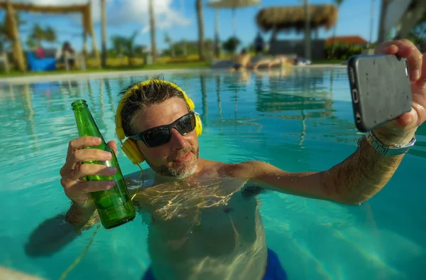 Joven feliz y atractivo hombre tomando foto selfie con el teléfono móvil bebiendo cerveza y escuchando música en la piscina tropical resort, piscina disfrutando de vacaciones de verano —  Fotos de Stock