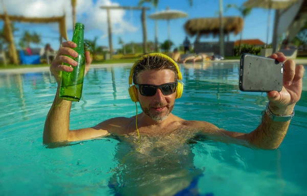 Joven feliz y atractivo hombre tomando foto selfie con el teléfono móvil bebiendo cerveza y escuchando música en la piscina tropical resort, piscina disfrutando de vacaciones de verano —  Fotos de Stock