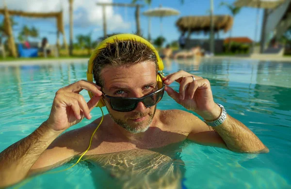 Hombre fresco y relajado de los años 30 disfrutando juguetón en la piscina tropical de lujo del resort escuchando música con auriculares sintiéndose complacido y fresco divirtiéndose en vacaciones de verano —  Fotos de Stock