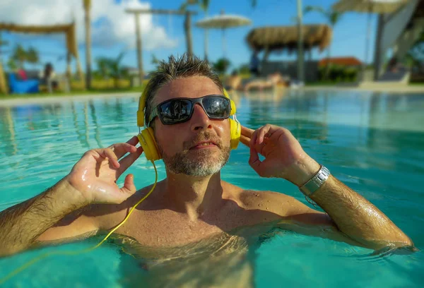 Hombre fresco y relajado de los años 30 disfrutando juguetón en la piscina tropical de lujo del resort escuchando música con auriculares sintiéndose complacido y fresco divirtiéndose en vacaciones de verano —  Fotos de Stock