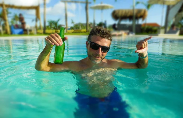 Joven hombre guapo y feliz en la piscina del hotel tropical beber botella de cerveza relajado y complacido disfrutar de vacaciones de verano viaje en turismo y viaje de vacaciones —  Fotos de Stock