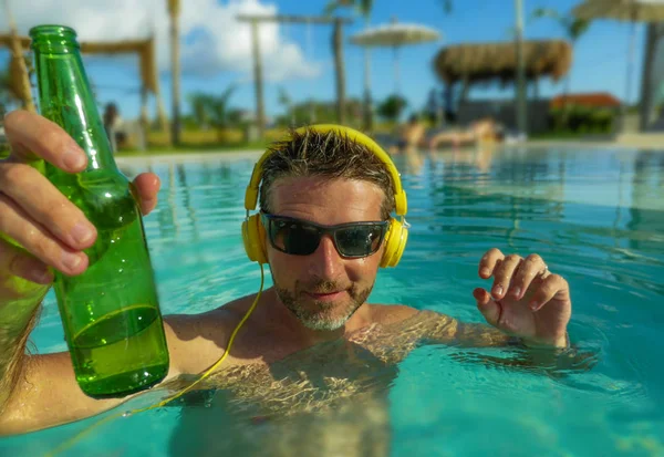 Joven hombre feliz y atractivo escuchando música con auriculares en la piscina del complejo tropical bebiendo cerveza disfrutando de un viaje de vacaciones de verano indulgente y de lujo —  Fotos de Stock