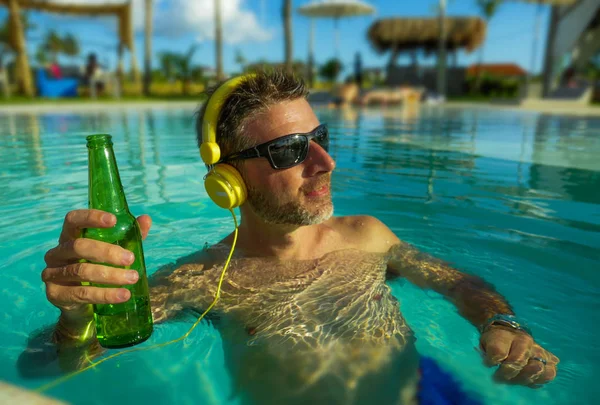 Jovem homem feliz e atraente ouvir música com fone de ouvido no resort tropical piscina beber cerveja desfrutando de férias de luxo e indulgente viagem de verão — Fotografia de Stock