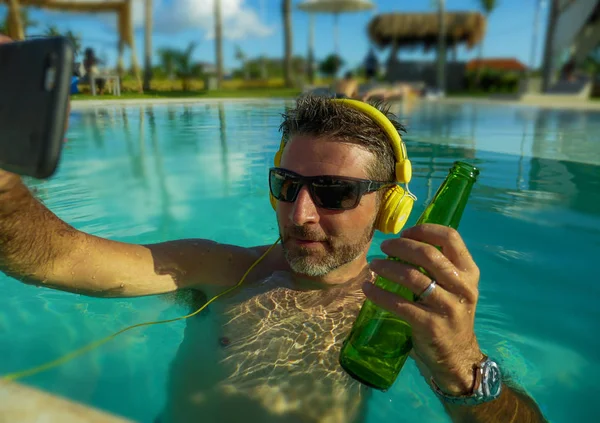 Joven feliz y atractivo hombre tomando foto selfie con el teléfono móvil bebiendo cerveza y escuchando música en la piscina tropical resort, piscina disfrutando de vacaciones de verano —  Fotos de Stock