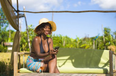 tropikal beach resort yüzme havuzunda Genç mutlu ve çekici siyah afro Amerikalı kadın bikini bardak şarap içme cep telefonunda internet sosyal medya kullanarak rahat